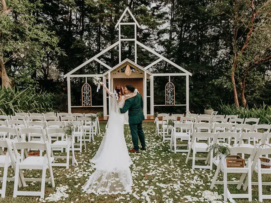 Novios frente al altar de la Fachada de la Iglesia, rodeados de sillas blancas y pétalos